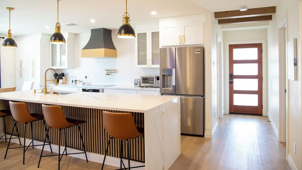 Una cocina moderna con una isla central, taburetes de bar, electrodomésticos de acero inoxidable, iluminación colgante y una puerta de madera.