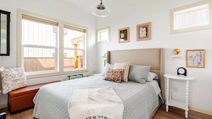 Quarto com uma cama grande, um banquinho, uma mesinha de cabeceira e uma janela com persianas.