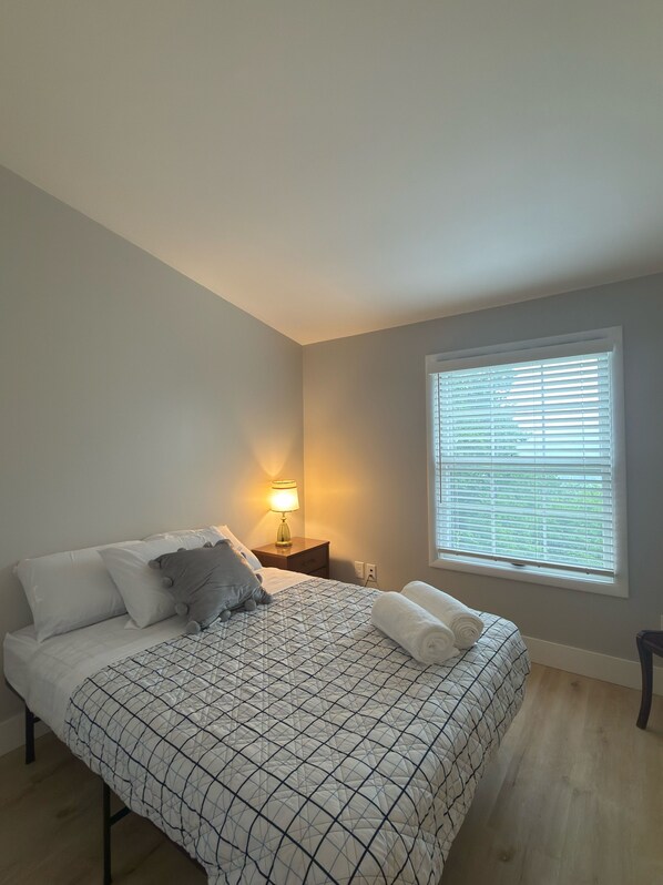 A bedroom with a bed, bedside lamp, window with blinds, and a view of greenery outside.