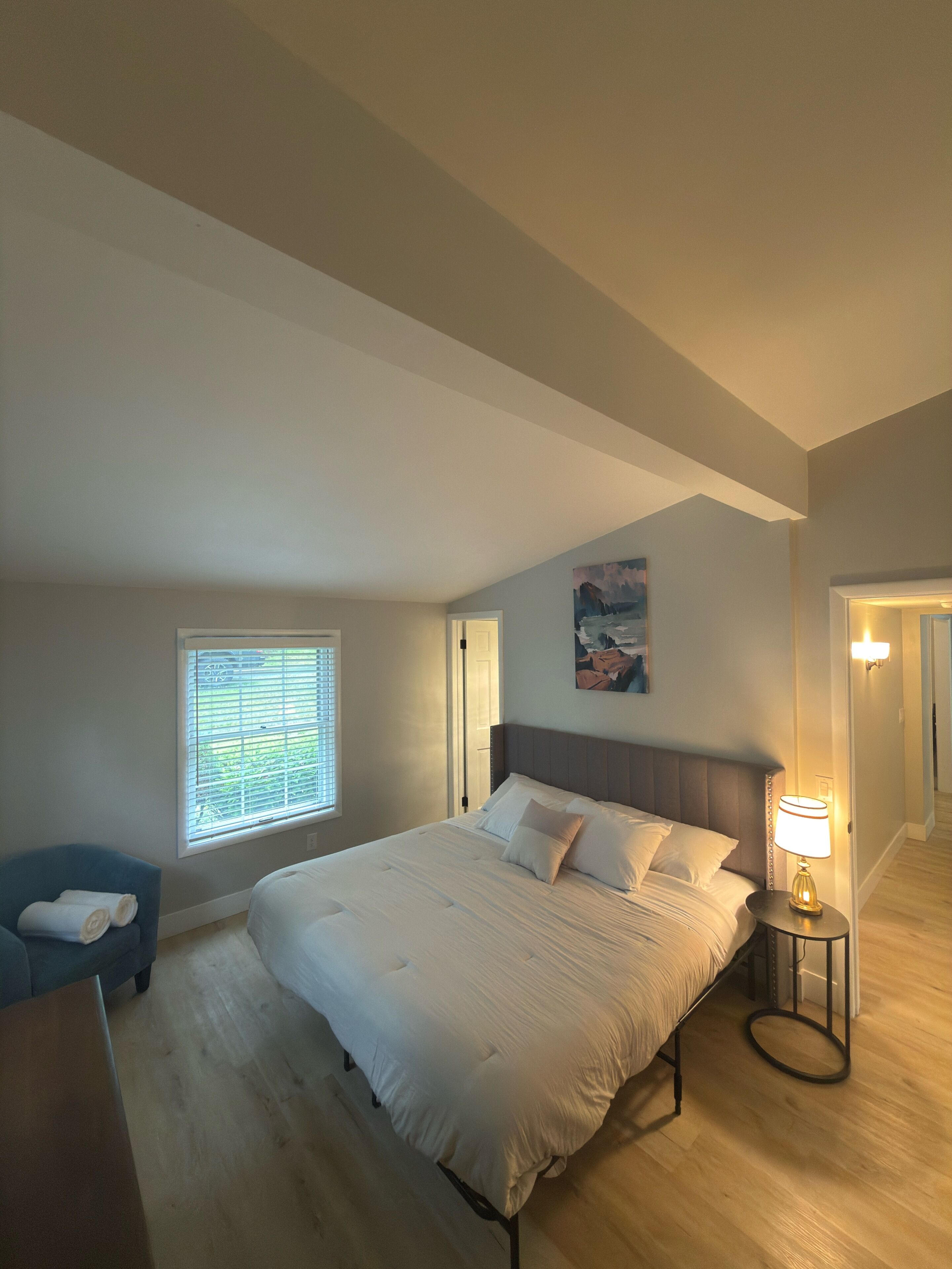 Une chambre avec un grand lit, une table de chevet avec une lampe, une fenêtre avec des stores, une chaise bleue et un tableau au mur.