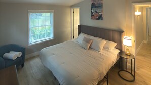 A bedroom with a large bed, a bedside table with a lamp, a window with blinds, a blue chair, and a painting on the wall. - Gorgeous, fully renovated 3-bedroom house with AC in tranquil La Vale (La Vale)