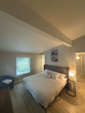 Une chambre avec un grand lit, une table de chevet avec une lampe, une fenêtre avec des stores, une chaise bleue et un tableau au mur.