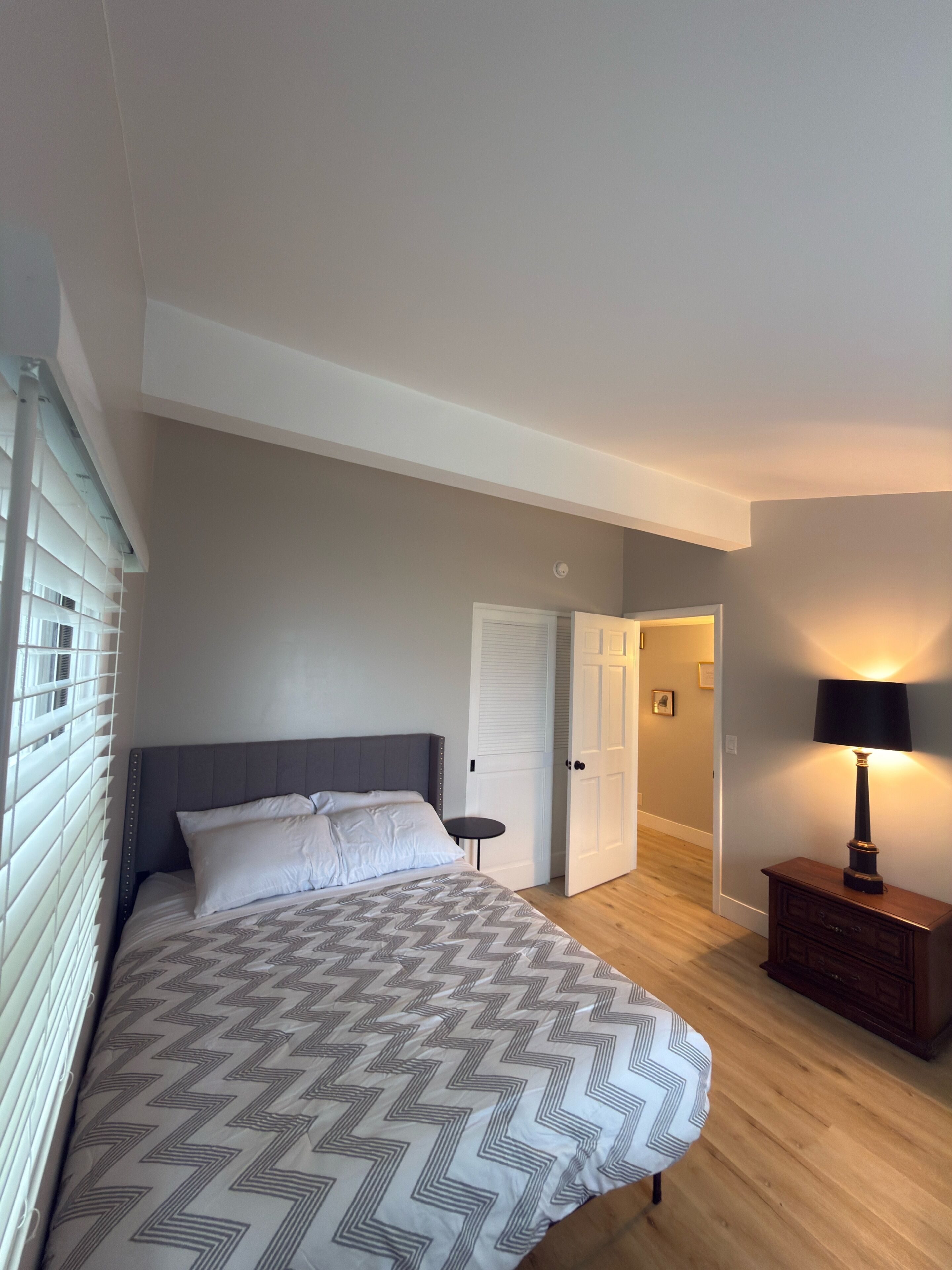 A bedroom with a bed, a bedside table, a lamp, a wardrobe, and a door leading to another room.
