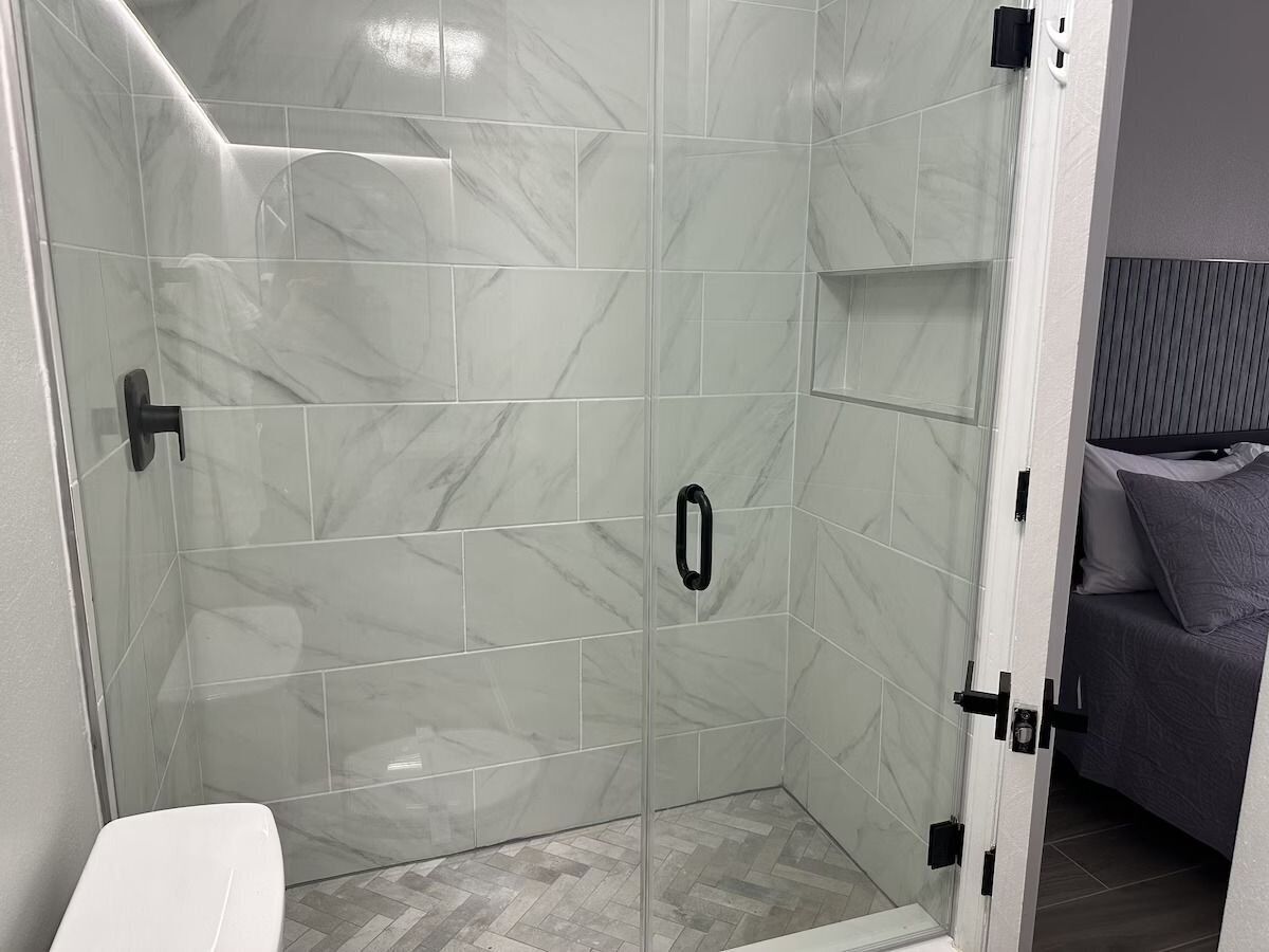 A shower with glass doors, a toilet, and a bed with a headboard in the adjacent room.
