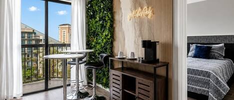 A modern hotel room with a balcony, a green wall, a coffee machine, and a bed with patterned bedding.