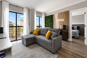 Un appartement moderne avec un canapé d’angle gris, un petit bureau, une machine à café, un lit avec une tête de lit grise et un mur vert agrémenté d’un jardin vertical.
