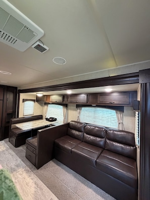 A compact RV interior with a kitchenette, a seating area with a sofa and a table, and a window with curtains.