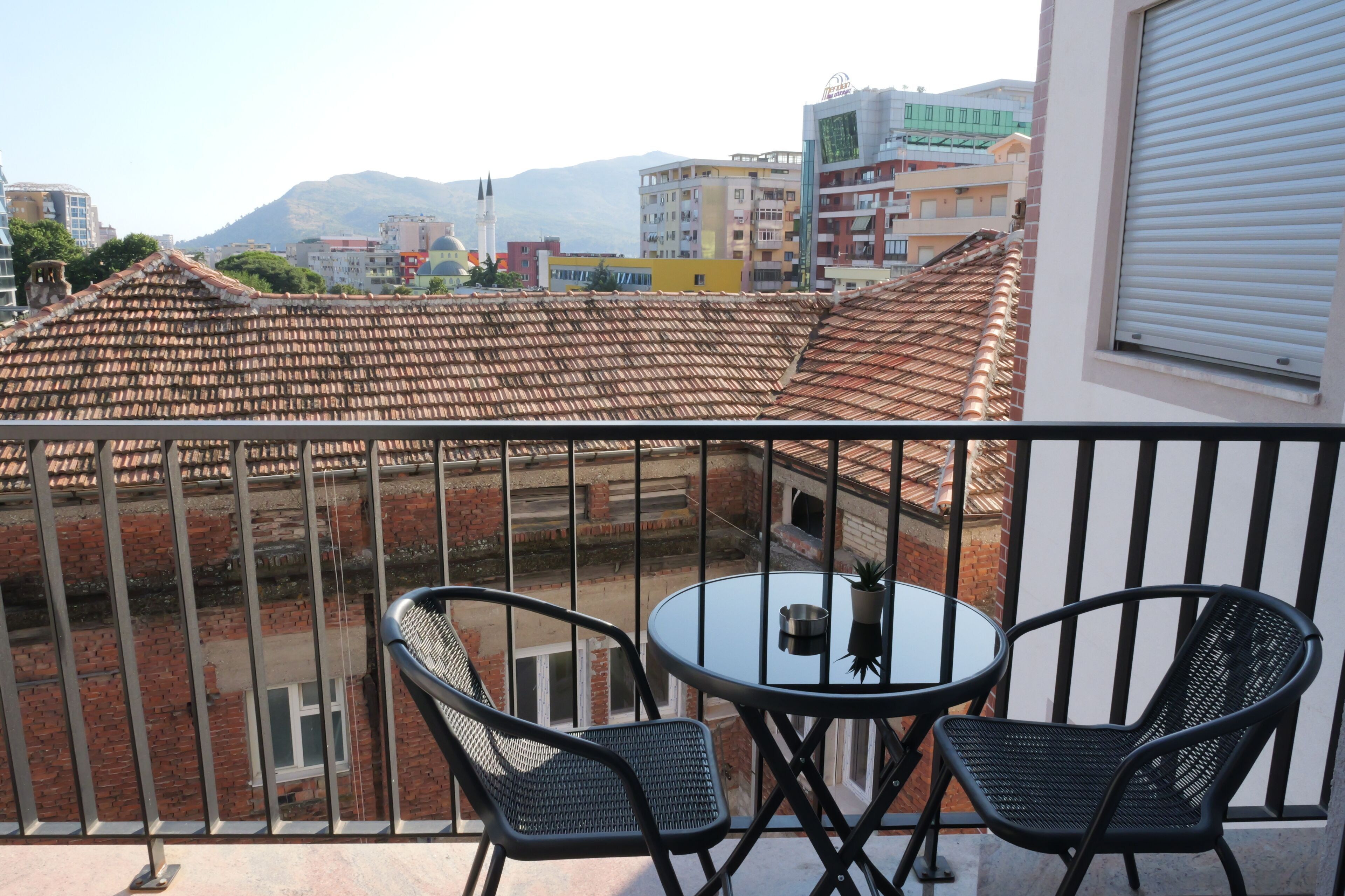 A balcony with a table and chairs, overlooking a cityscape with buildings and a mountain in the background.