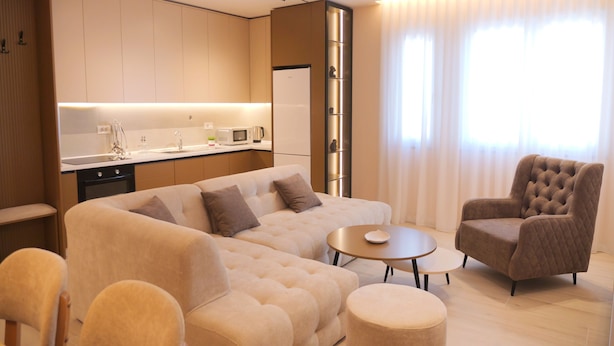 A modern living room with a sofa, armchair, and ottoman, a kitchen area with appliances, and large windows with curtains. - Horizont Premium Apart-Hotel (Shkodër)