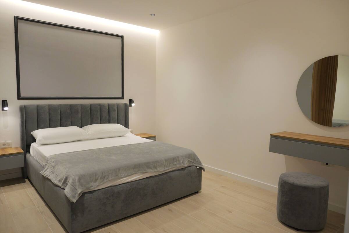 A modern bedroom with a bed, bedside tables, a nightstand, a round mirror, and a small stool.