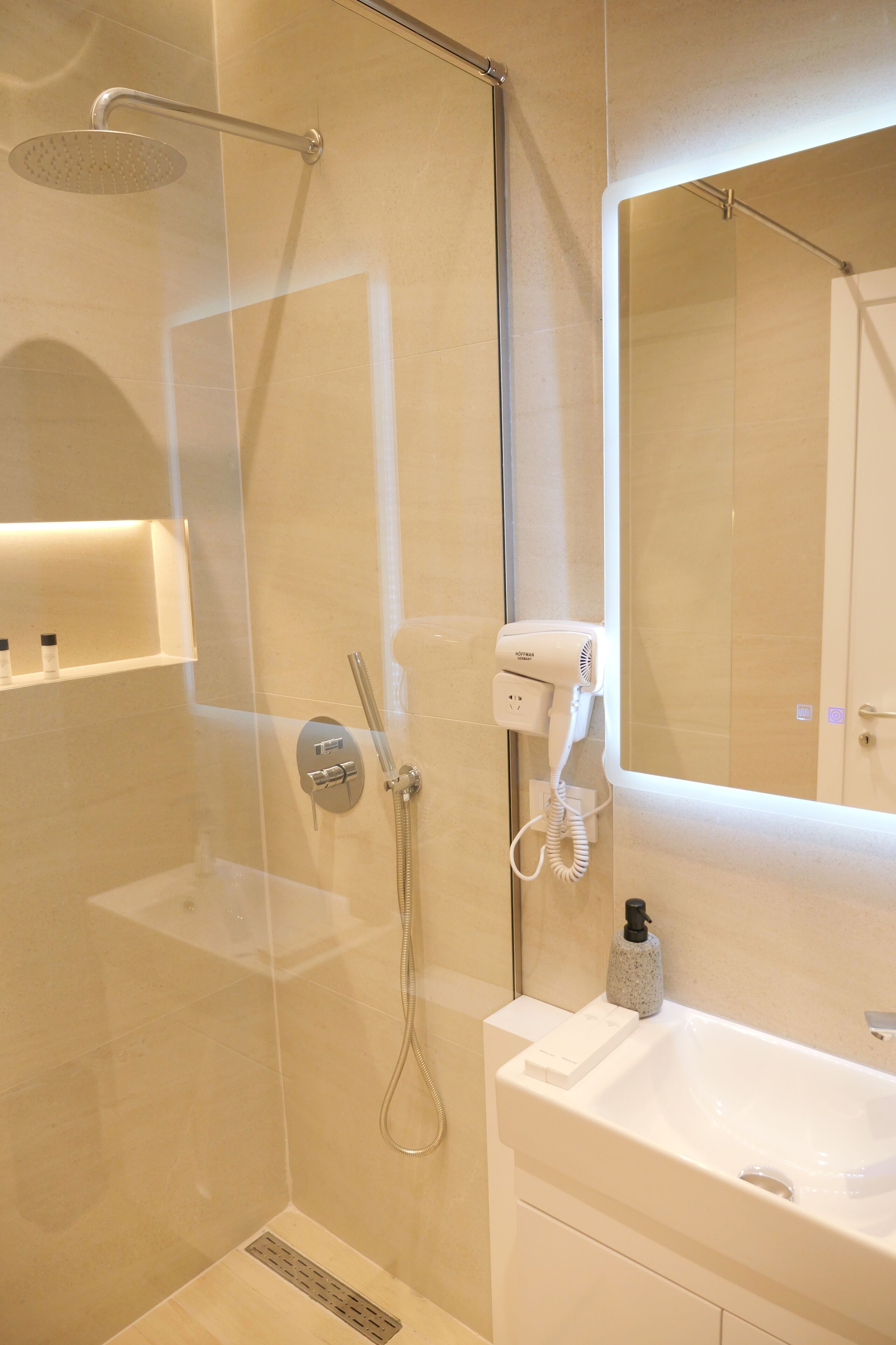 A modern bathroom with a glass-enclosed shower, a handheld showerhead, a wall-mounted hairdryer, and a white sink with a mirror above it.