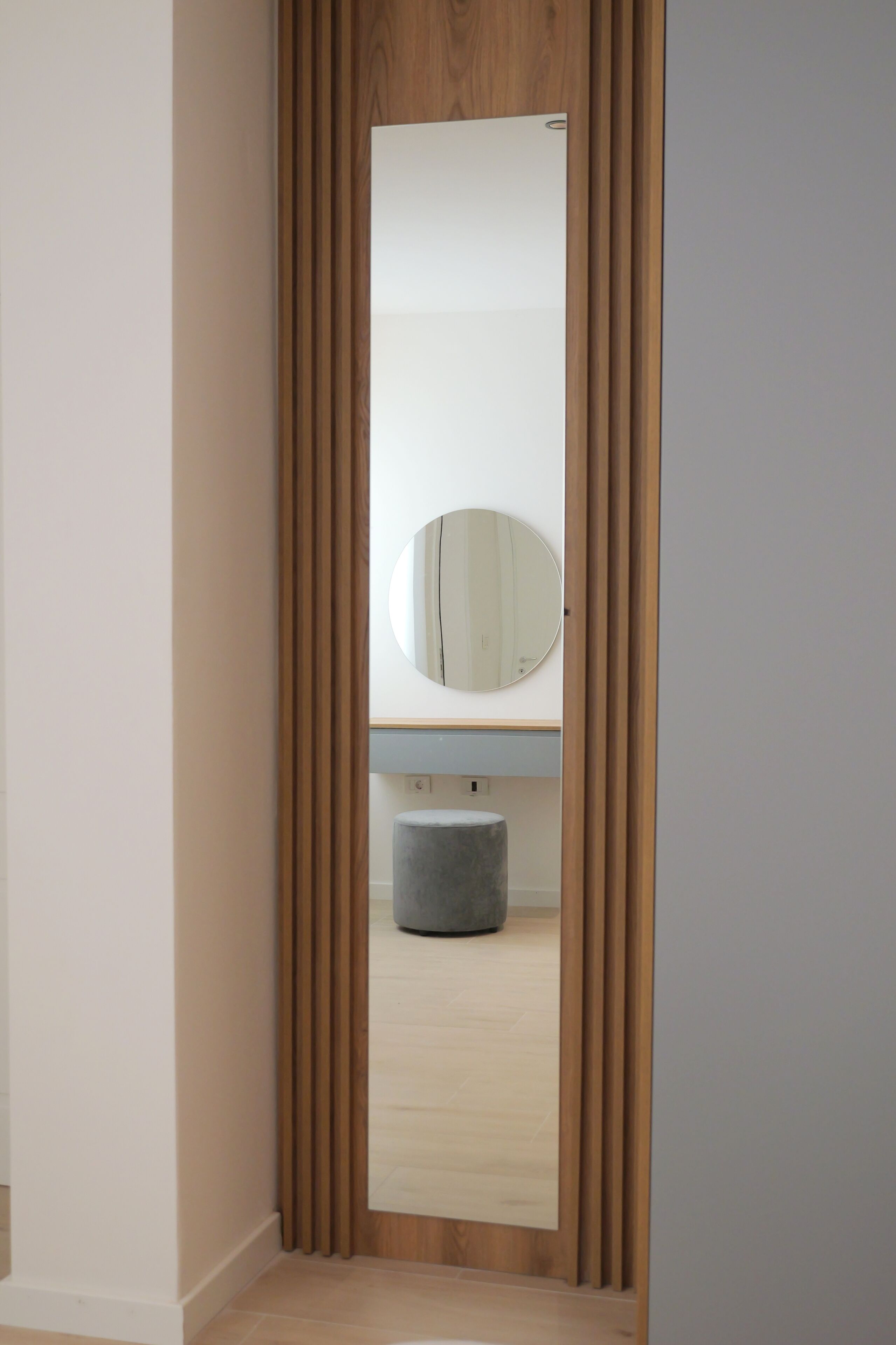 Un couloir étroit aux murs recouverts de lambris en bois, un miroir rond accroché au mur et un pouf gris dans la pièce.