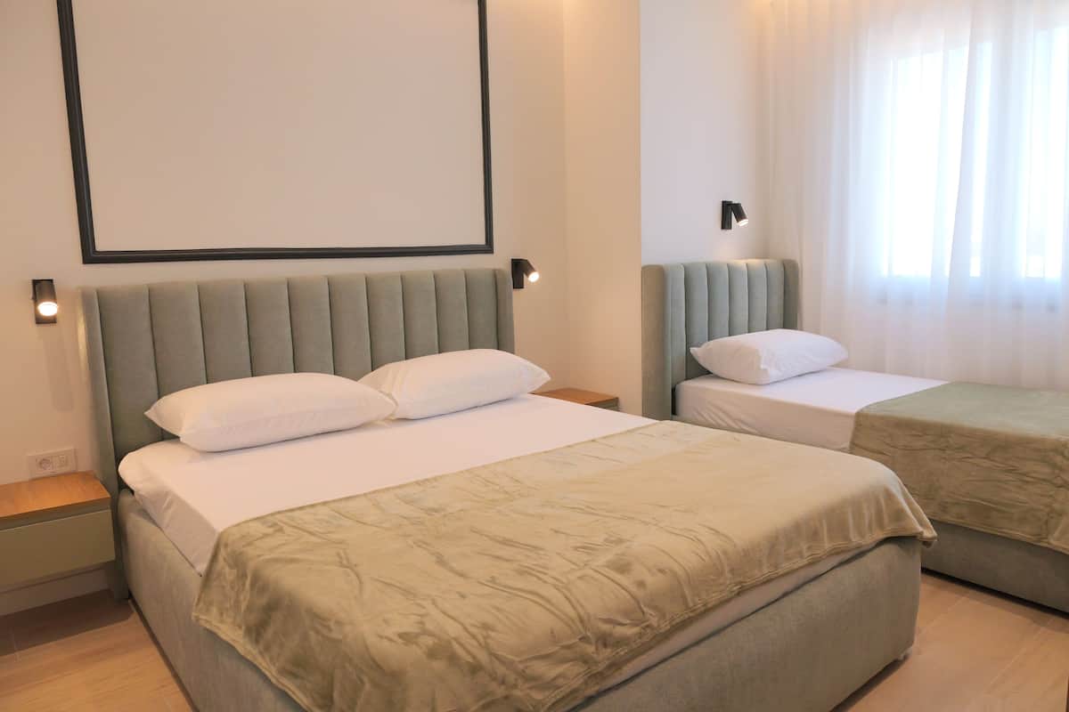 A hotel room with a large bed, a smaller bed, a nightstand, a wall-mounted light, and a window with sheer curtains.