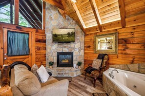 Un interior de cabaña acogedor con chimenea de piedra, televisor montado, puerta de madera y una bañera.