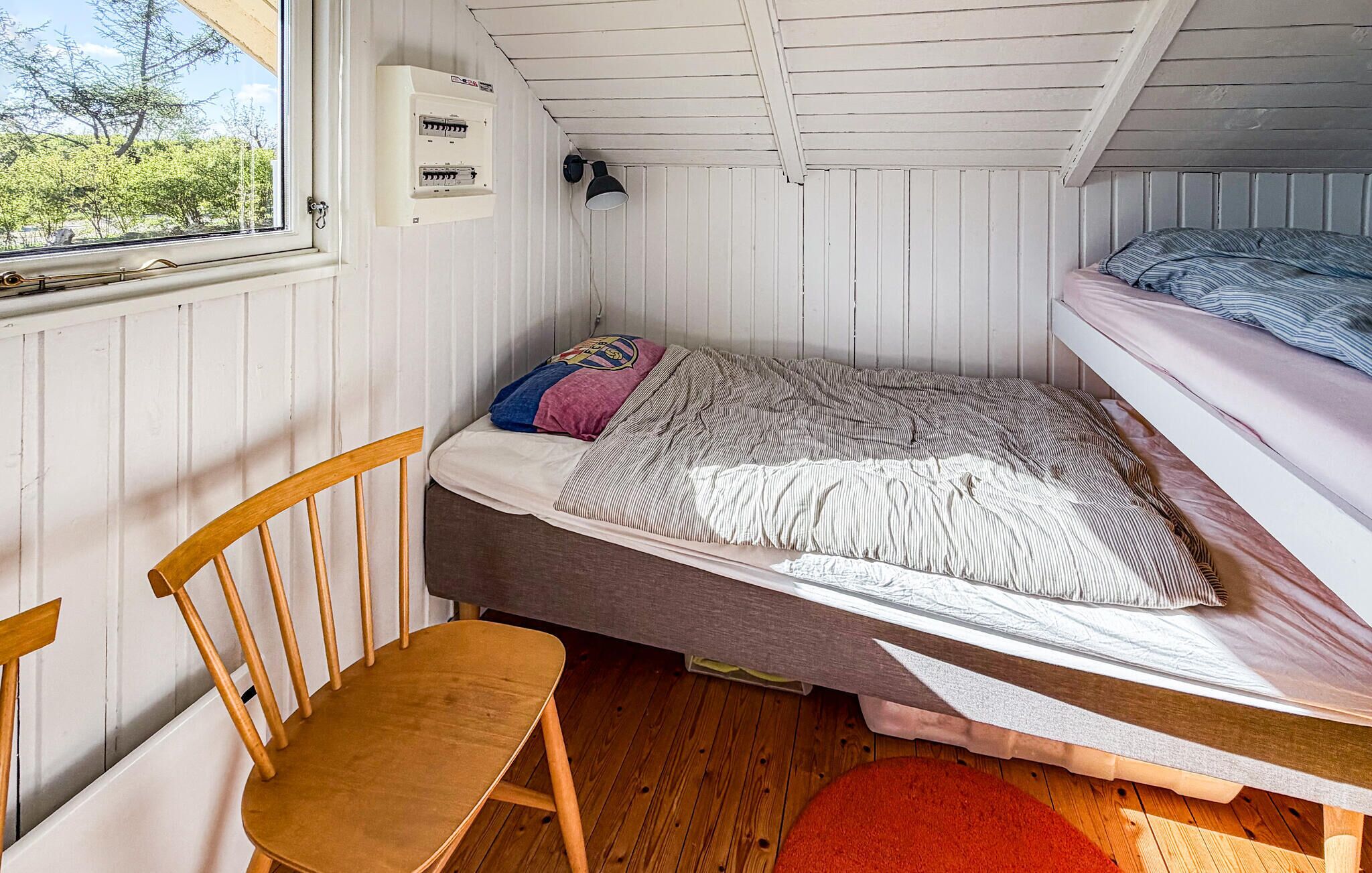 A small, single-bed room with a wooden chair, a window, and a wall-mounted electrical panel.