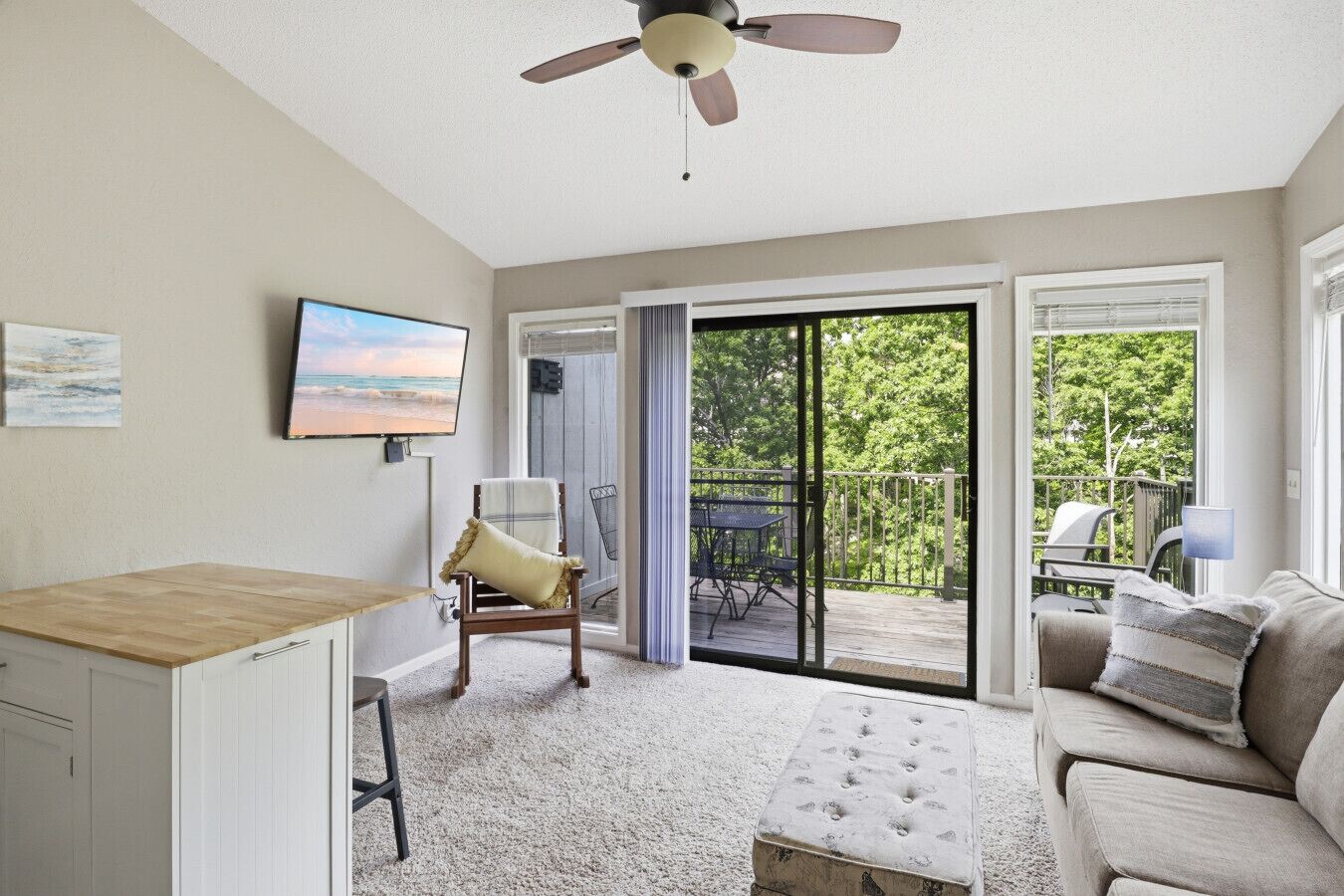 A room with a ceiling fan, a TV, a wooden desk, a cushioned chair, a sofa, and a sliding glass door leading to an outdoor deck.