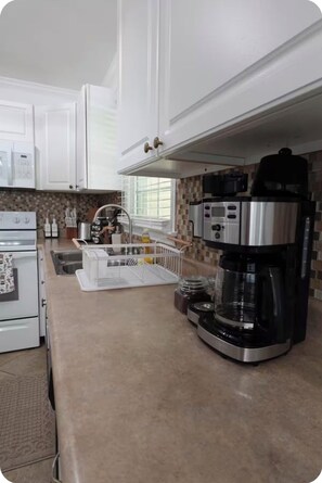 A kitchen with white cabinets, a microwave, a stove, a coffee maker, and a window with blinds. - Relaxing 3BR Near RAFB with AC & WiFi (Warner Robins)