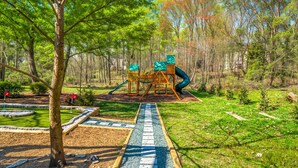Un parque infantil con un tobogán y estructuras de escalada, rodeado de un jardín ajardinado con un sendero pavimentado.