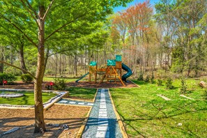 A playground with a slide and climbing structures, surrounded by a landscaped garden with a paved path. - 5BR Movie Theater, Hot Tub, Mini Golf, Game Room! (McGaheysville)
