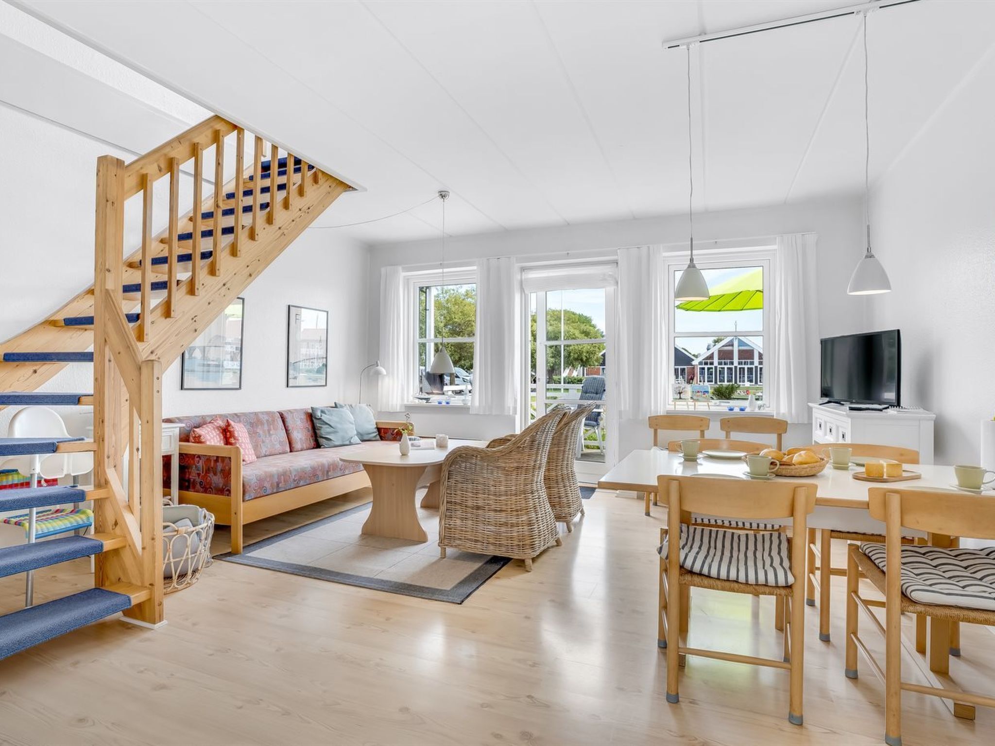 Un vaste espace de vie comprenant un escalier en bois, un canapé, une table à manger, des chaises en osier et un téléviseur.