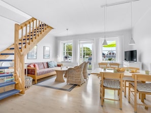 Ein geräumiger Wohnbereich mit einer Holztreppe, einer Couch, einem Esstisch, Korbstühlen und einem Fernseher.
