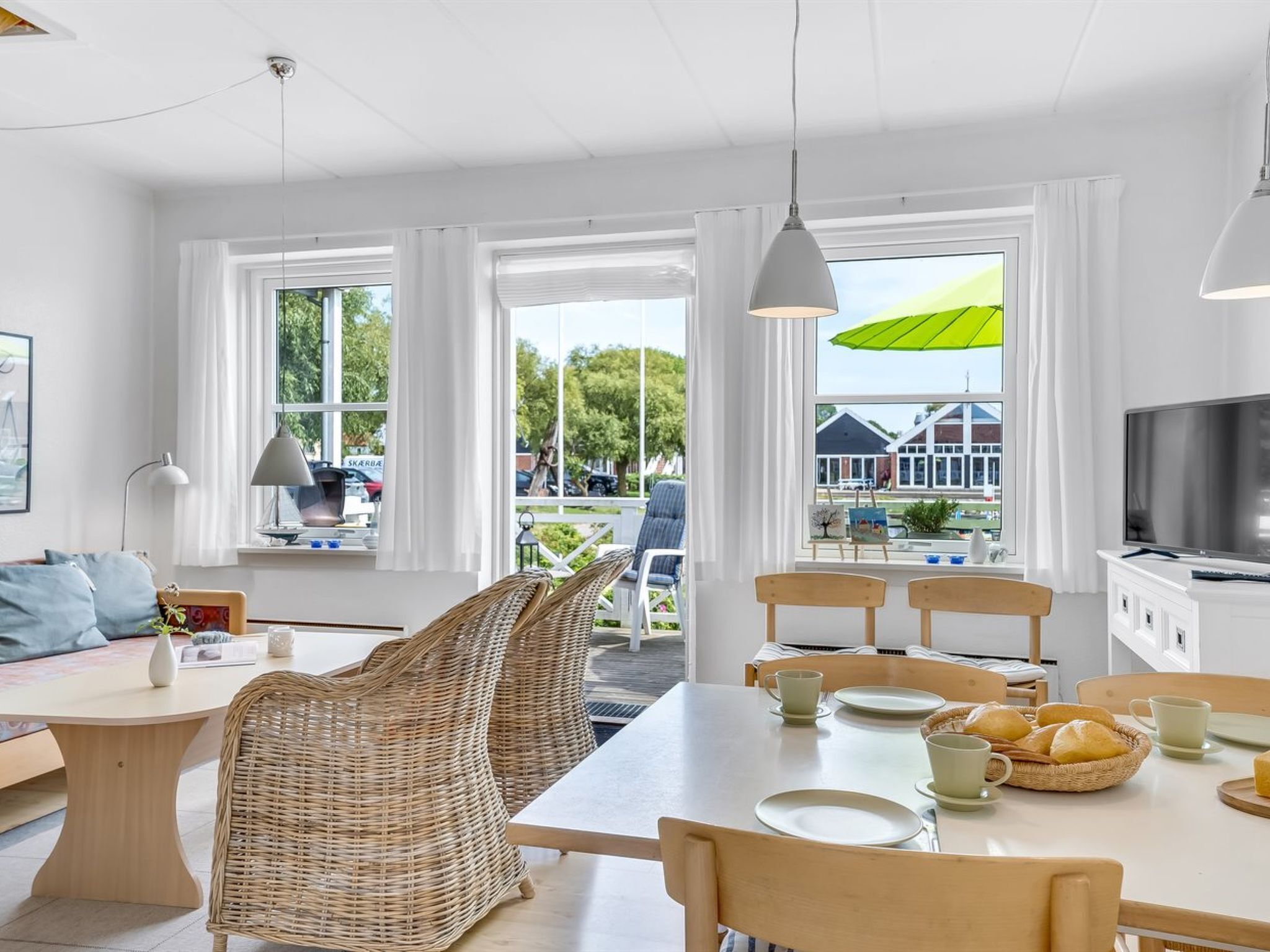 Une pièce moderne et bien éclairée, avec une table à manger dressée pour deux personnes, des chaises en osier, un canapé et un téléviseur.