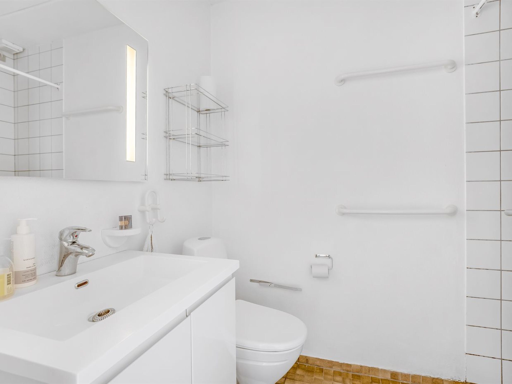 Ein modernes Badezimmer mit Waschbecken, Spiegel und Toilette in Weiß. Zwei Handtuchhalter, ein Duschbereich und ein kleines Regal mit Pflegeprodukten.