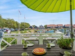 Eine Holzterrasse mit Tisch, Stühlen, einem grünen Sonnenschirm und Blick auf eine Marina mit Booten und Gebäuden.