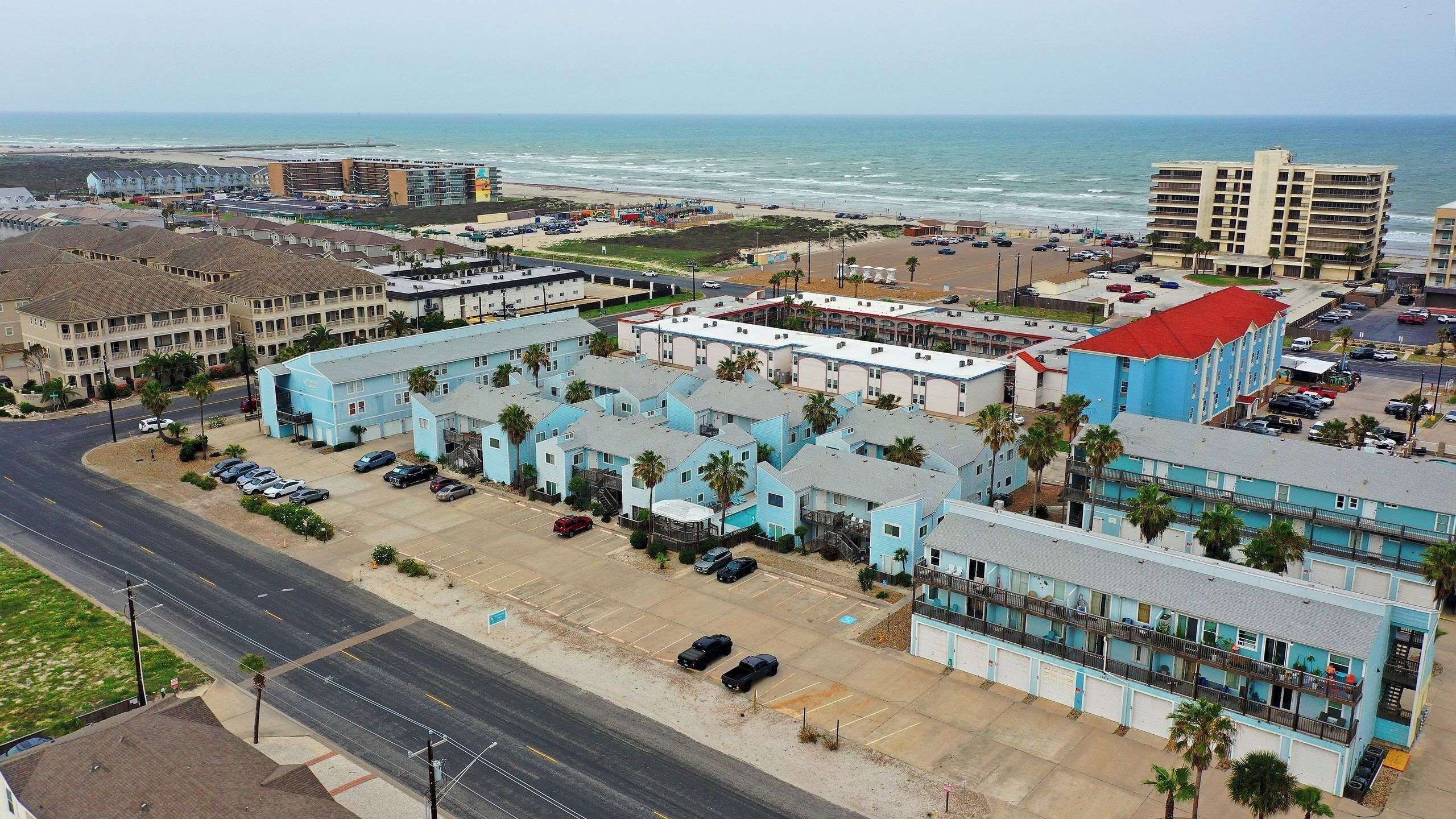 A coastal hotel with multiple-story buildings, a parking lot, and a view of the beach and ocean.