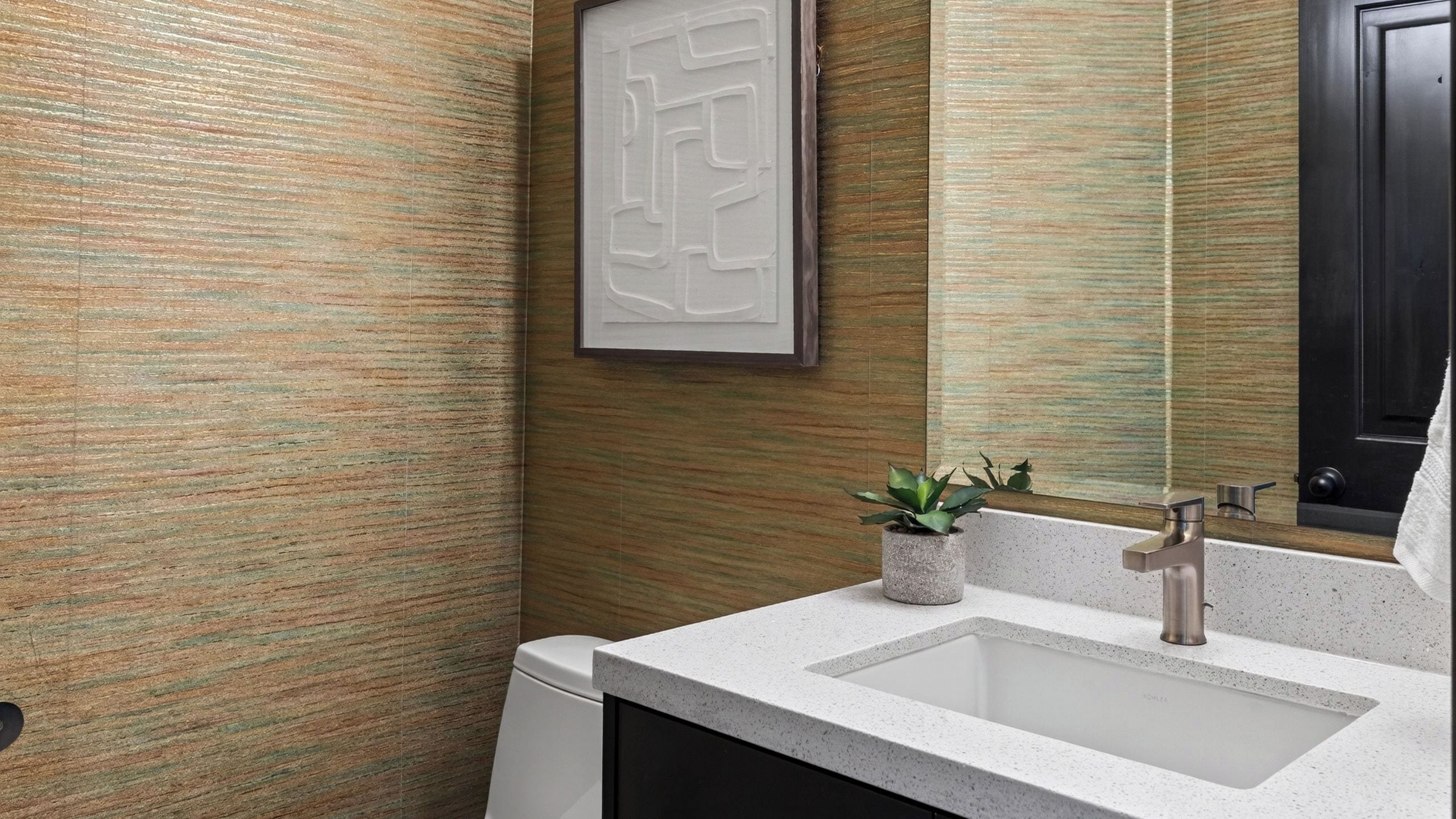 Un baño con pared texturizada, un lavabo con grifo moderno, una pequeña planta en maceta y una obra de arte enmarcadas.