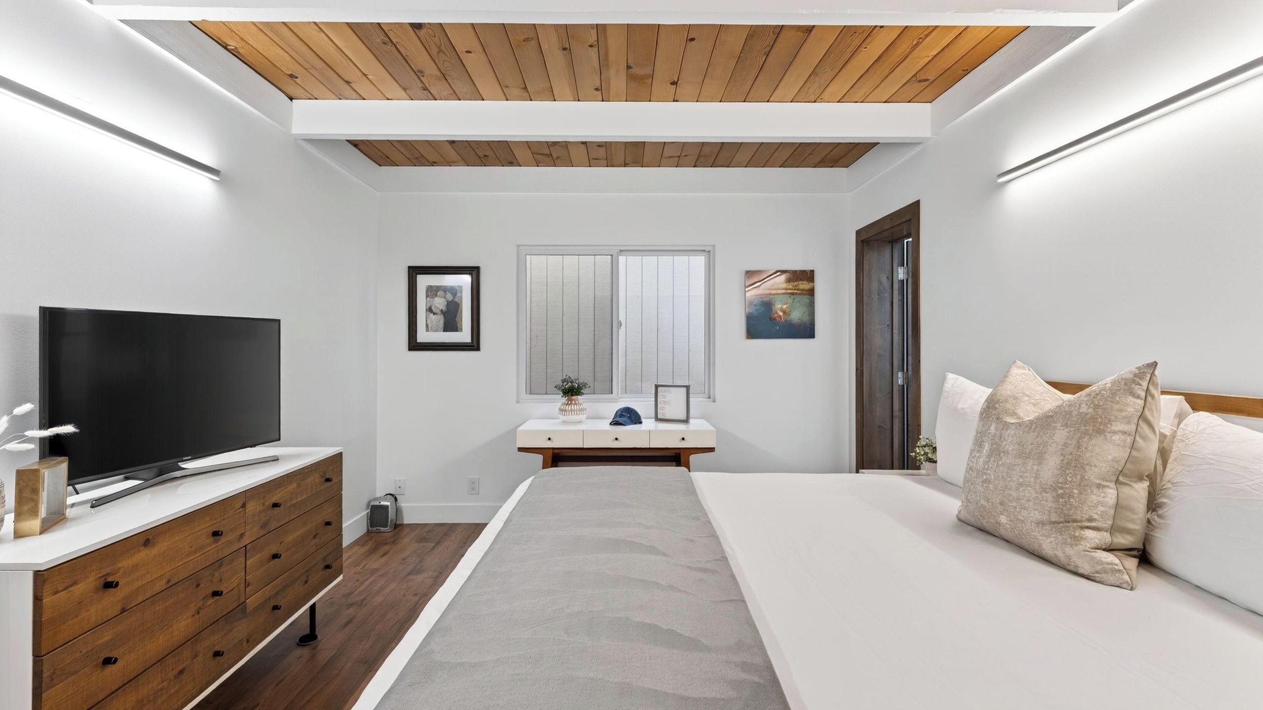 A modern bedroom with a TV, wooden dresser, bedside table, and a bed with pillows.