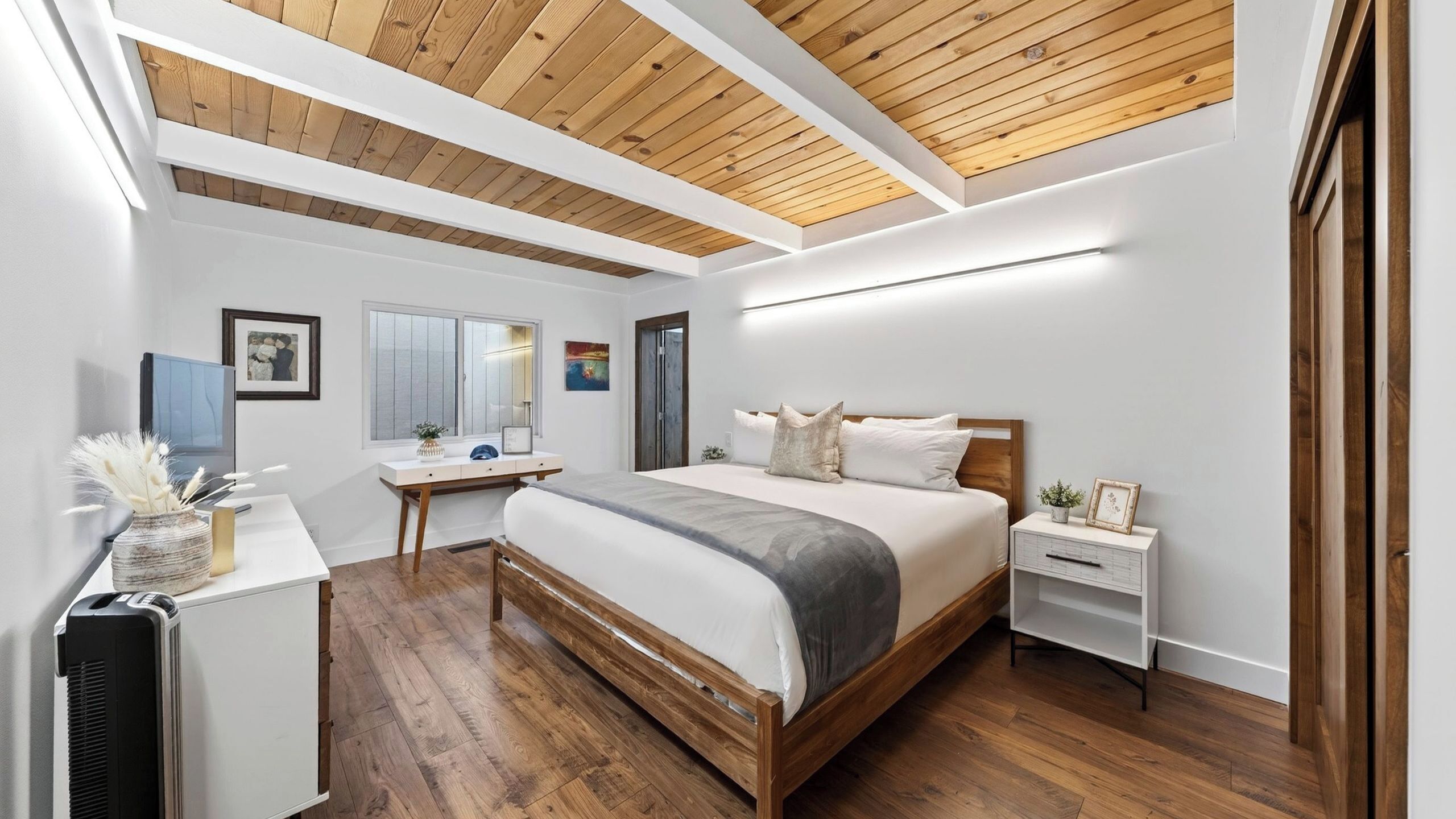 A bedroom with a wooden ceiling, a bed with white bedding, a desk, and a nightstand.