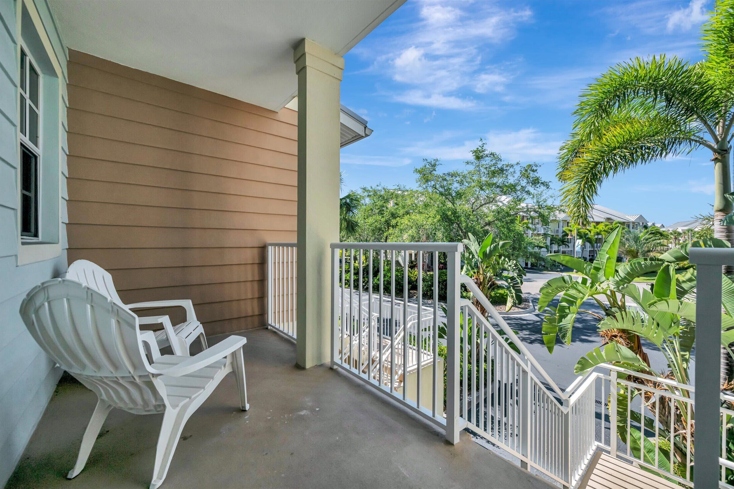 A balcony with a white chair, a railing, and a view of a street with houses and greenery.