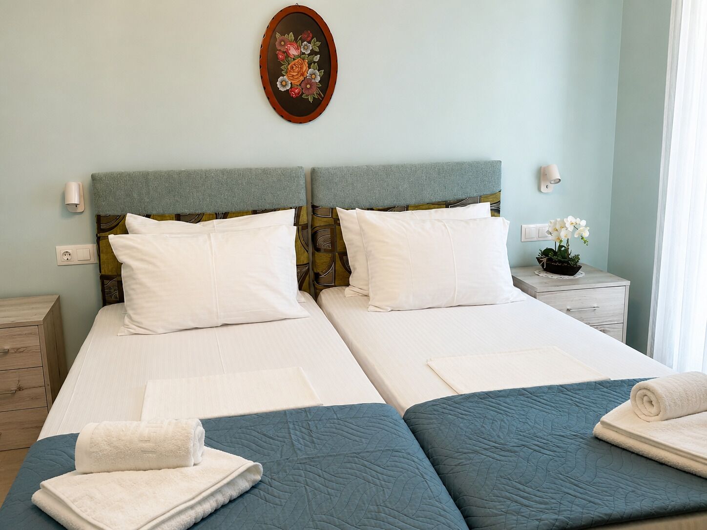 A double bed with two pillows, a bedside table with a lamp, a decorative wall piece, and a window with curtains.