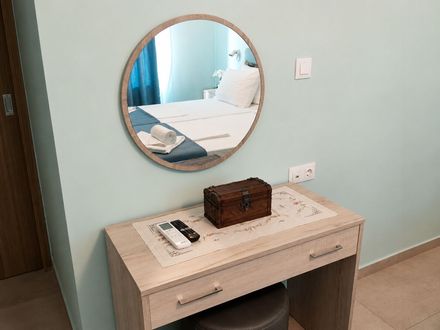 Una habitación de hotel con una cama, una mesita de noche con un control remoto, una pequeña caja de madera y un espejo redondo en la pared.