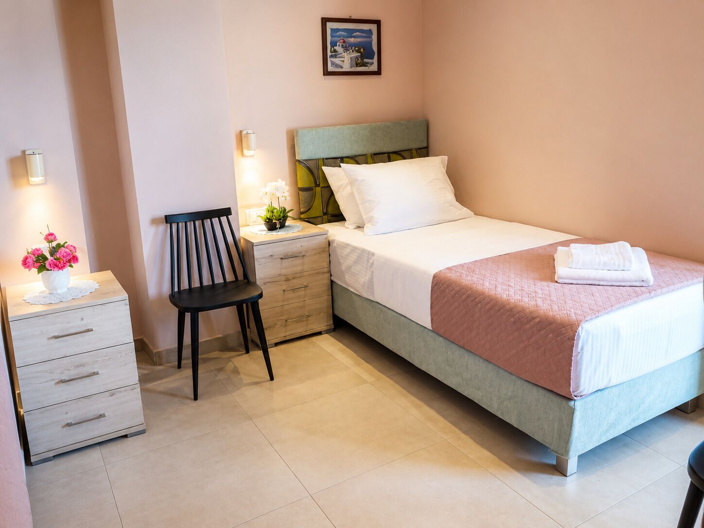 A neatly arranged bedroom with a bed, a bedside table with a lamp, a chair, and a small desk with drawers.