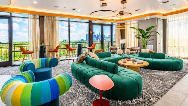 Ein modernes Wohnzimmer mit einem grünen Ecksofa, bunten Sesseln und einer roten Stehlampe.
