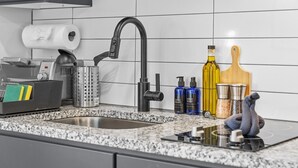Eine Küche mit Spüle aus Edelstahl, einer Spülmittel-Zapfsäule, einer Handseifen-Zapfsäule, einer Olivenölflasche, einem Holzschneidebrett und Salz- und Pfefferstreuern.