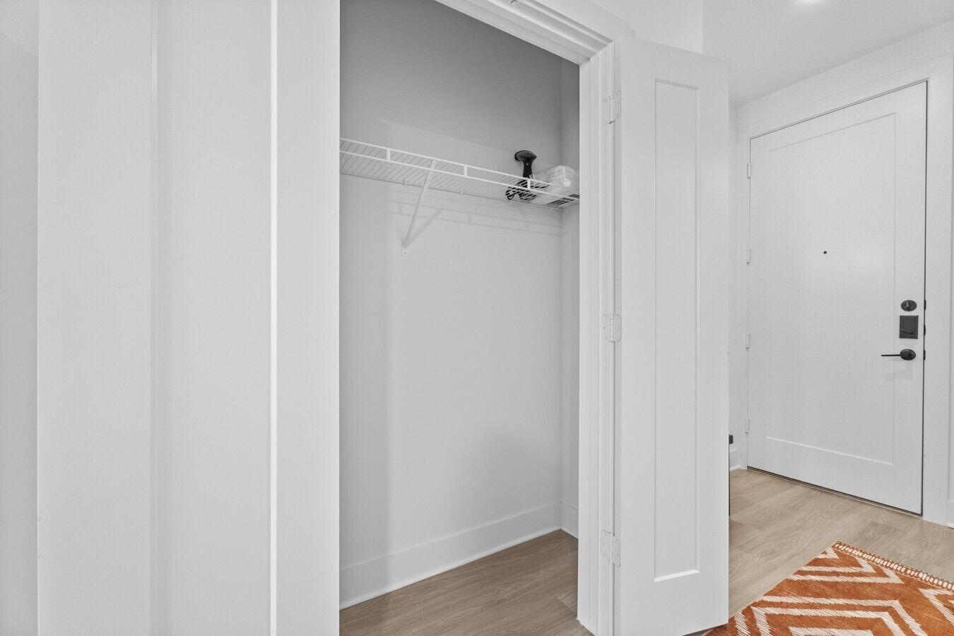 Un couloir intérieur aux murs blancs, doté d’un plancher en bois, d’une porte blanche et d’un placard avec une étagère en métal.