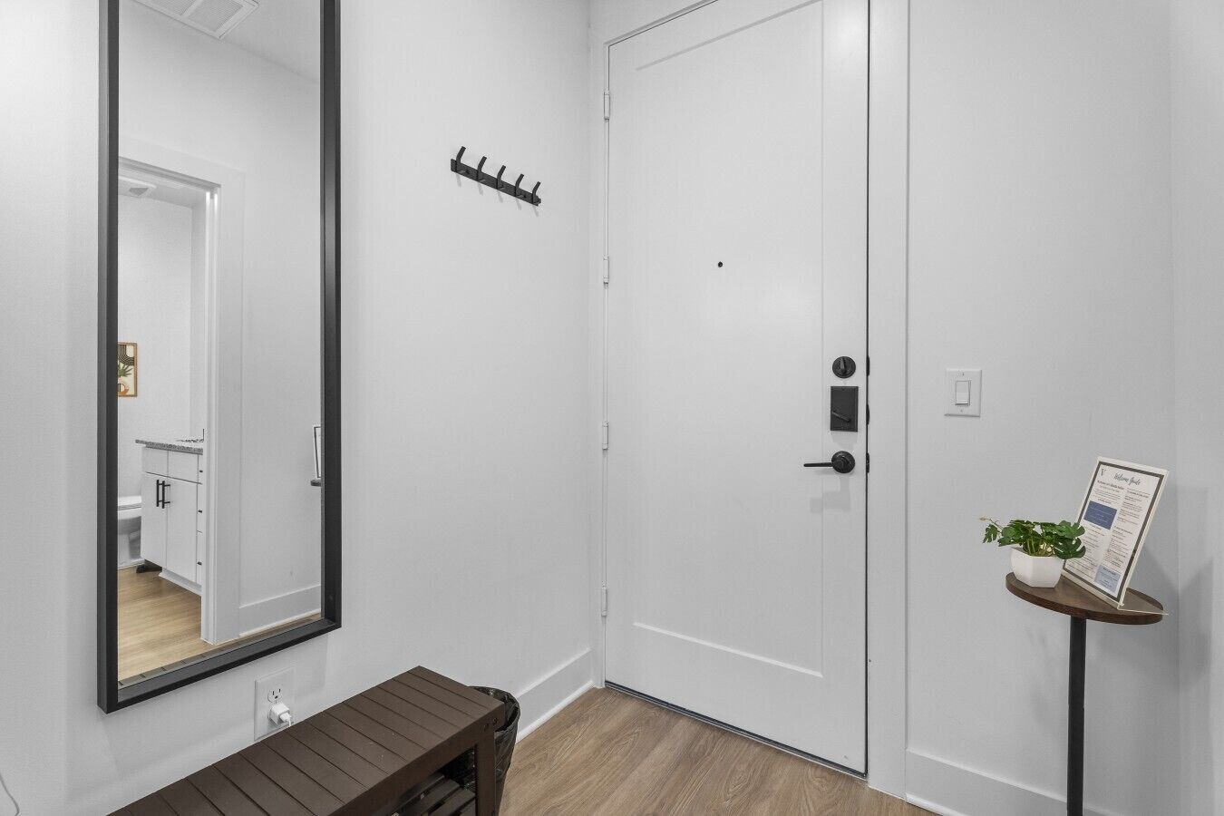 A white door with a lock, a coat rack, a small table with a plant, and a mirror reflecting a bathroom.