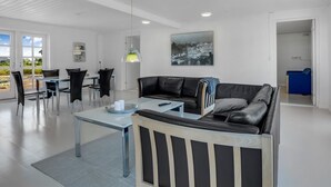 Ein modernes Wohnzimmer mit einem Glastisch, einer schwarzen Ledersofa, einer Essecke mit weißem Tisch und Stühlen sowie einem Stadtbild an der Wand.
