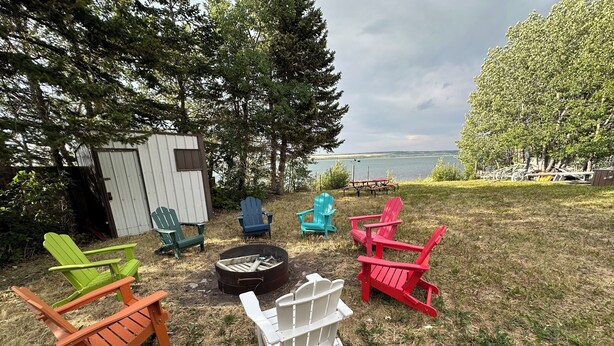 A backyard with Adirondack chairs in various colors, a fire pit, and a view of a lake. - Montana Moon on the Lake "White Hawk" RV #3 (Babb)