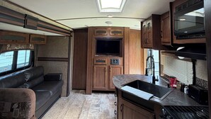 A compact RV kitchen with a built-in stove, a sink, and a television. - Montana Moon on the Lake "White Hawk" RV #3 (Babb)