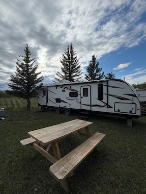 A recreational vehicle with a 'White Hawk' logo, a picnic table, and two large evergreen trees in the background. - Montana Moon on the Lake "White Hawk" RV #3 (Babb)