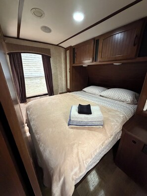 A compact RV bedroom with a bed, a small bedside table, a window with curtains, and a ceiling light. - Montana Moon on the Lake "White Hawk" RV #3 (Babb)