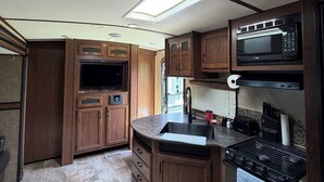 A compact RV kitchen with wooden cabinets, a microwave, a stove, and a sink. - Montana Moon on the Lake "White Hawk" RV #3 (Babb)