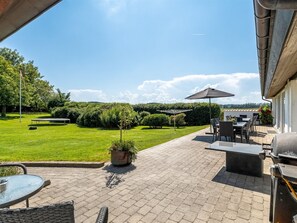 Eine Terrassenfläche mit Gartenmöbeln, einem Grill und Blick auf einen gepflegten Rasen und Bäume.