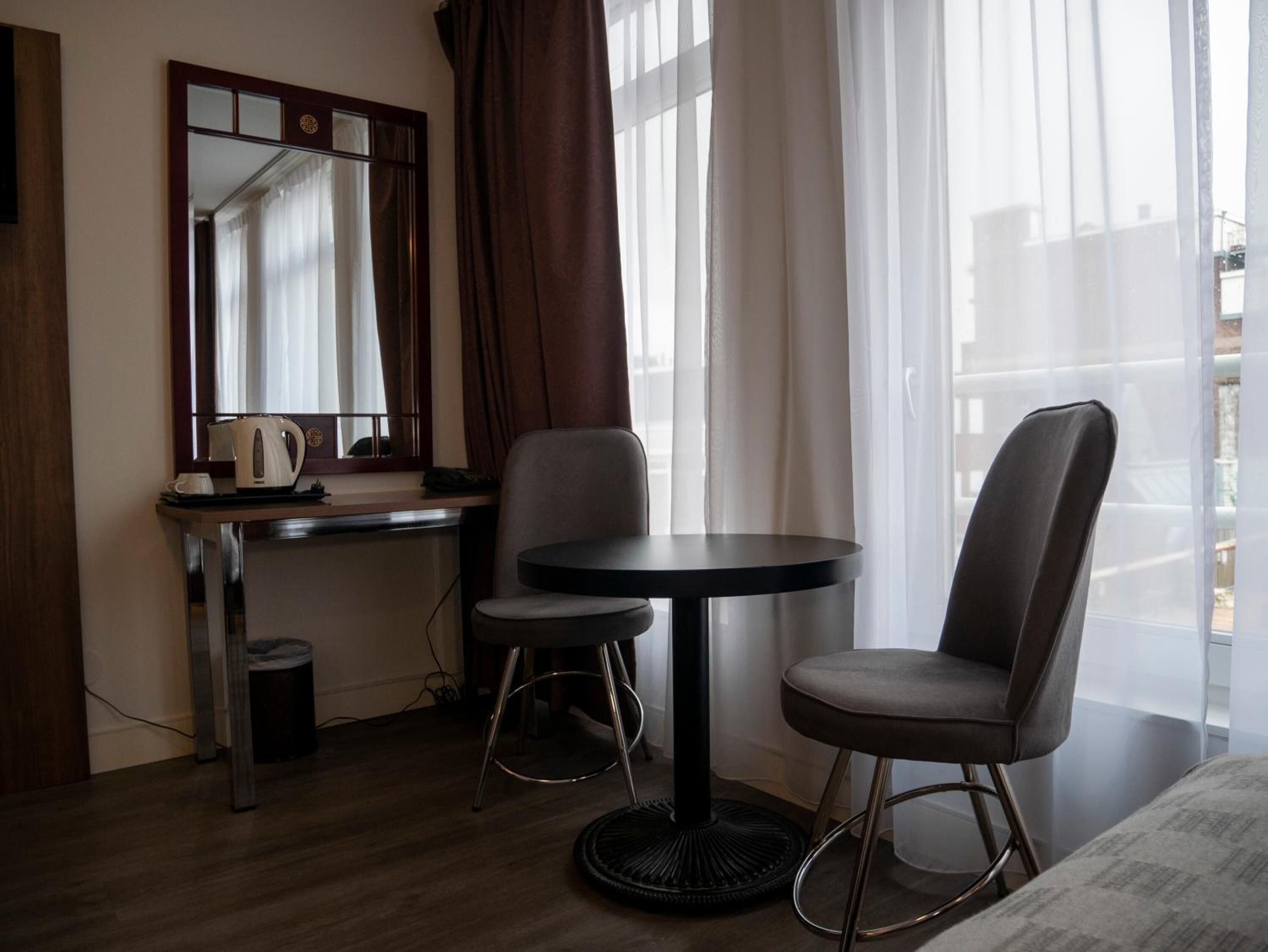 Een hotelkamer met een bureau, twee stoelen, een ronde tafel, een spiegel en een raam met gordijnen.