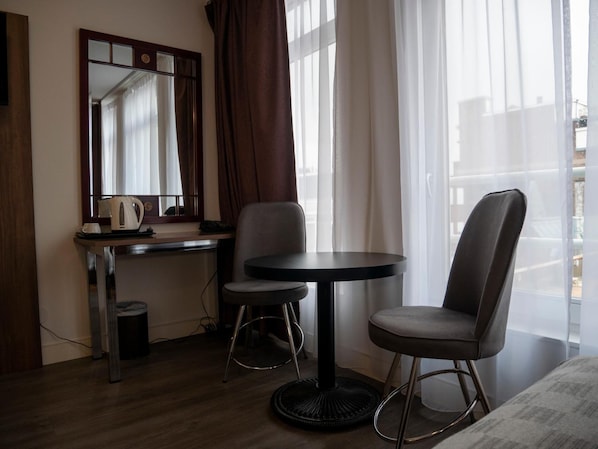 Quarto de hotel com escrivaninha, duas cadeiras, mesa redonda, espelho e janela com cortinas.
