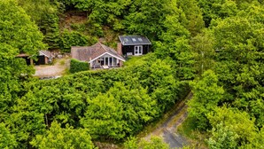 Uma casa rodeada por floresta densa, com uma entrada de automóveis visível a levar até à porta.